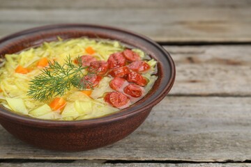 Bowl of delicious sauerkraut soup with smoked sausages and dill on wooden table, closeup. Space for text