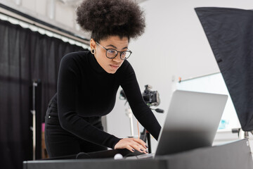 concentrated african american content producer using laptop in photo studio on blurred foreground.