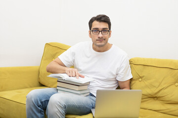 Confused man looking at the screen of laptop and sitting on the couch with a lot of books. Online learning and home education concept