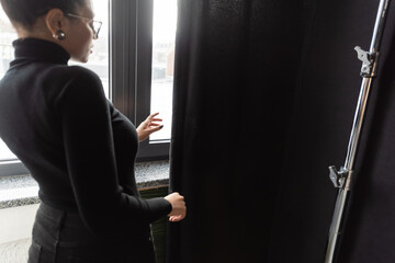 african american content manager in turtleneck standing near window and black curtain in photo studio.