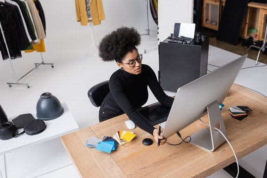 high angle view of african american retoucher adjusting computer monitor near graphic tablet and color swatches on desk in photo studio.