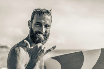 Surfer happy with surf surfing smiling doing hawaiian Shaka Brah or Hang Loose during surf session in ocean waves on beach vacation -  Friendly greeting in surfer culture - Black and white editing