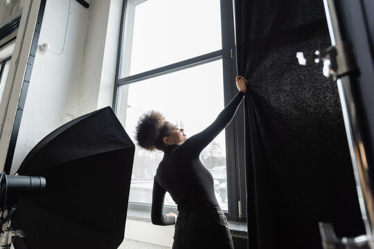 african american content manager in black turtleneck pulling curtain near window near softbox reflector in photo studio.