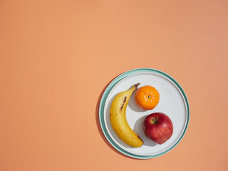 Fruits on a plate. Whole unpeeled banana, apple and tangerine on a white plate on another turquoise plate. View from above on orange background with copy space.