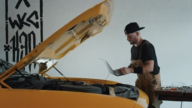 Lateral medium full shot of tattooed auto mechanic checking car engine under opened yellow motor hood, connected to laptop mechanic holding in his hands. White wall with signs in backgroundLateral med