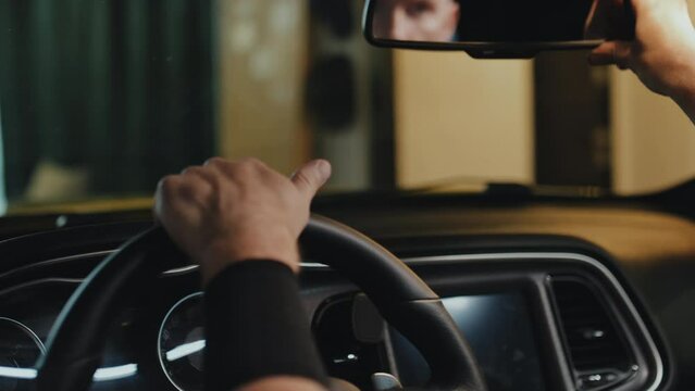 Loose close up of male tattooed hands on steering wheel, right hand moving rearview mirror, his face reflected in it, garage interior in background, cropped image