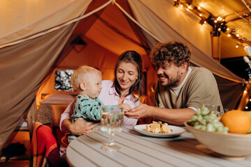 Happy family with lovely baby have dinner and spend time together in glamping on summer evening near cozy bonfire. Luxury camping tent for outdoor recreation and recreation. Lifestyle concept