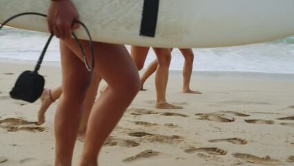 Legs of surfer girls walking along beach carrying surfboards at sunrise. Female surfers feet walk on beach along ocean at dawn. Slim fit legs of young surfing women walking in ocean tide