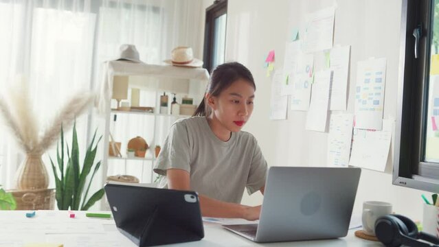 Young Asia girl IT development programmer typing on keyboard coding programming fixing data code on laptop computer screen follow code plan in tablet and paperwork on table workroom at house office.