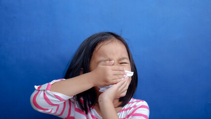 A cold girl blows her nose in a white rag on blue background. Influenza or Rhinitis snot runny nose stuffy nose. Allergy young asian girl holdingwhite  handkerchief. Medical concept.