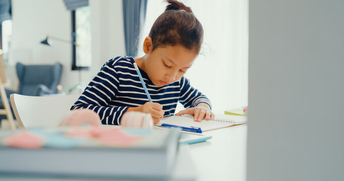 Asian toddler girl with sweater sit in front of desk with notepad use pencil focus on write notebook do homework from online learning course on the weekend at home. Distance online learning concept.