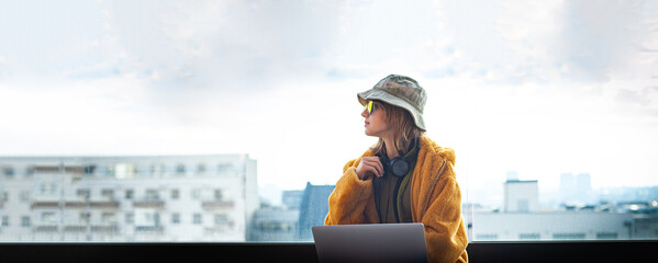 Hipster teenage girl sitting with laptop on terrace with view on city. Young happy woman in hat...