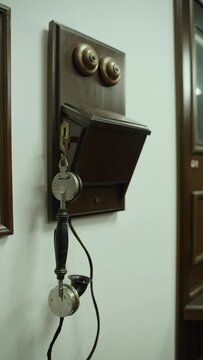 Antique wall-mounted citophone with wooden panel with bell hangs on wall, vertical shot. Early 20th century telephone receiver with wooden handle and cord hangs on metal eyelet on base, nobody.