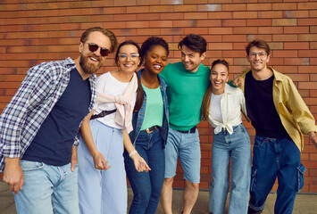 Hanging out with friends. Portrait of carefree multiracial friends in casual clothes on background of brick wall. Cheerful men and women having fun standing in row hugging and smiling at camera.