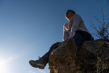high rock, woman seated with legs together, short-haired person in her forties, non-binary looking towards the sky.