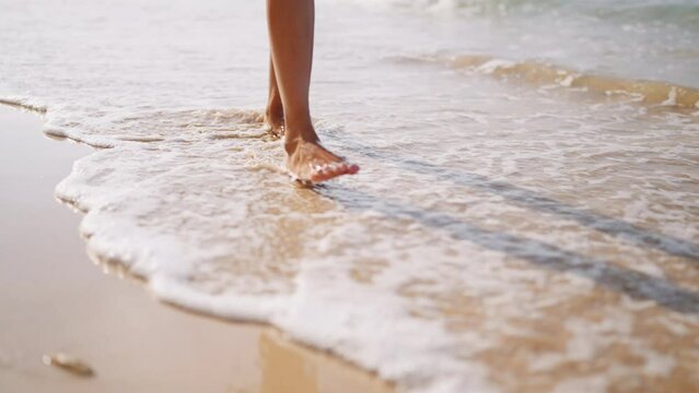 Black woman feet walking on sandy ocean beach at exotic island along sea water waves. Dark skin female legs go by surf line, touch white foam. Bipoc girl relaxing on tropical travel at luxury resort.