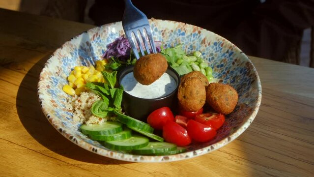 Close up of fork dipping a falafel in creamy sauce. Chickpea falafel salad with vegetables, quinoa and Edamame beans with Zaziki sauce on a wooden table. Vegetarian vegan food. High quality 4k footage