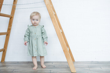 Portrait of serious little blond barefoot girl with blue eyes wearing green dress, standing under wooden stepladder.