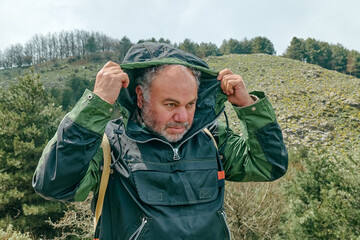 Mature bearded traveler man hiking in mountain forest, wearing jacket with hood to protect himself from the wind in the mountain valley.