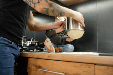 Cropped photo of man shaking, turning over metal bowl of kneader machine, pulling out dough on table near little boy.