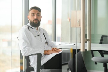 Naklejka na ściany i meble Portrait of arabic doctor handsome young man in workwear posing at modern clinic, sitting at workdesk