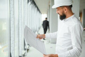 Naklejka na ściany i meble Serious busy young attractive arabic industrial engineer in hard hat with project drawings in factory interior. Professional and industry, builder architect job, worker checking work at plant indoor