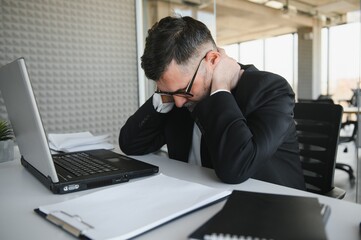 Worried businessman in dark suit sitting at office desk full with books and papers being overloaded with work.