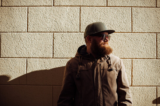 Portrait of handsome bearded ginger man with sunglasses and green baseball flat cap, against yellow (beige) wall. Monochrome street style picture.