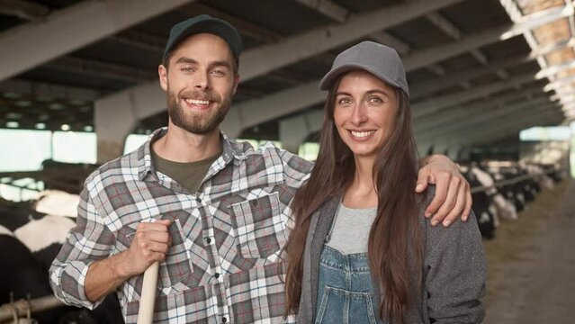 Portrait of Caucasian couple with cap standing in barn with cows. Attractive man hugging his woman and smiling on camera. Young village people caring cattle together. Concept of couple farmers.