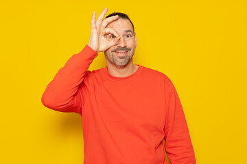 Bearded hispanic man in his 40s wearing a beige turtleneck making an ok gesture looking through fingers, isolated on yellow studio background.