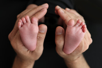 A mother hold the feet of a newborn child in a black blanket on a Black background. The feet of a newborn in the hands of parents. Studio macro photo legs, toes, feet and heels of a newborn.