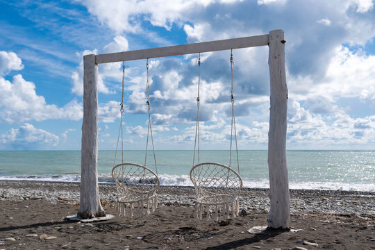 Wicker chair hammock. Comfortable hanging wicker white chair on the seashore. A cozy place to relax with a sea view. Boho style hammock chair hanging on a tree. The concept of outdoor recreation near 