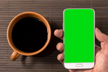 A person holding a cell phone with a green chroma screen next to a cup of coffee on a wooden...