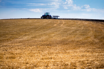 Obraz premium A modern blue tractor with an attached reversible plow with a husking roller is plowing a field on which the spring grain crop has just been harvested. Midsummer in central Ukraine.