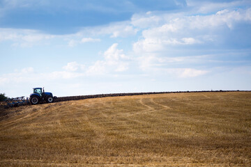 A modern blue tractor with an attached reversible plow with a husking roller is plowing a field on which the spring grain crop has just been harvested. Midsummer in central Ukraine.