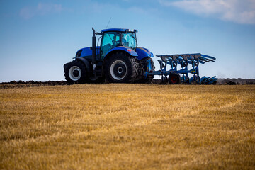 A modern blue tractor with an attached reversible plow with a husking roller is plowing a field on which the spring grain crop has just been harvested. Midsummer in central Ukraine.