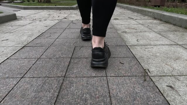Female feet in black sneakers walk along the paths and sidewalks of city streets and parks