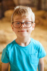a boy with glasses in a blue T-shirt with a strange expression on his face stands against the background of the barn from the inside