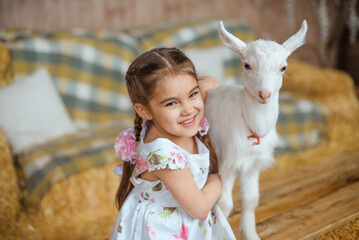 girl is hugging a white baby goat. She is laughing, happy, and wearing a summer cotton dress with braided hair and pink ribbons. They are in a rural barn surrounded by hay and both are looking at the