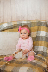 A little infant girl sits on a checkered blanket, softly caressing a fluffy rabbit with her tiny hands.