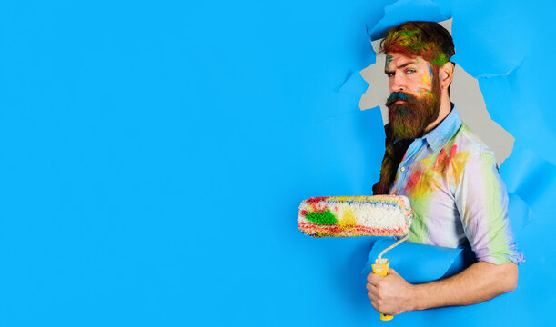 Painter man. Bearded painter with painting roller looking through hole in paper. Handsome bearded worker with paint roller. Professional painter in dirty dye shirt. Serious painter man. Copy space.