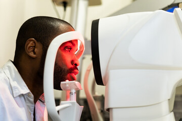 A dark-skinned man is undergoing a vision test using a corneal topographer to examine the cornea....