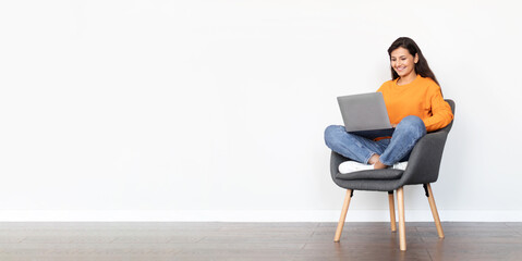 Positive middle eastern young lady using laptop, sitting in armchair