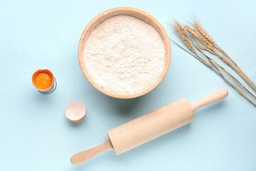 Bowl with flour, wheat ears, cracked egg and rolling pin on blue background