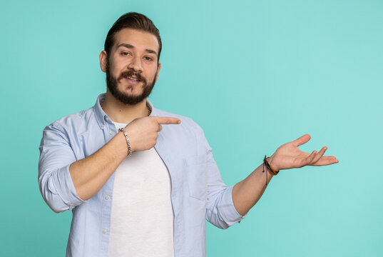 Young lebanese man showing thumbs up and pointing empty place, advertising area for commercial text, copy space for goods promotion. Bearded arabian guy isolated on blue studio background, indoors