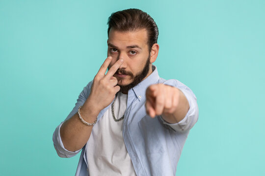 I am watching you. Young confident lebanese man pointing at his eyes and camera, show I am watching you gesture spying on someone. Handsome bearded arabian guy isolated alone on blue studio background