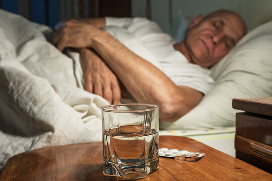 Lonely Elderly Sick Man Sleeping In Bed With Pills And Water Glass On Wooden Nightstand Foreground. Senior People Health Care Concept.