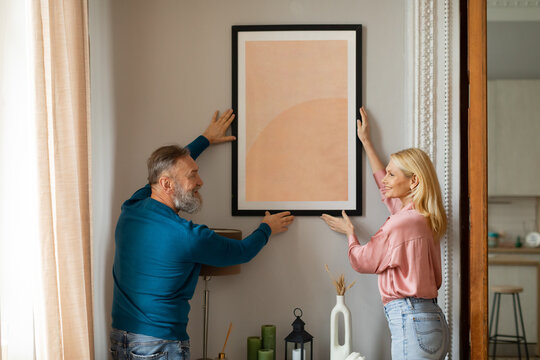 Happy Mature Couple Hanging Picture In Frame On Wall Indoors