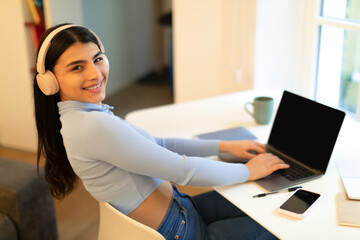 Happy mexican student lady sitting at desk, using wireless headphones and laptop with blank screen, mockup