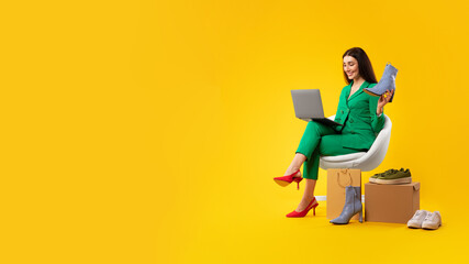 Stylish lady using laptop and holding elegant shoes, consulting clients online, sitting on yellow background, copy space
